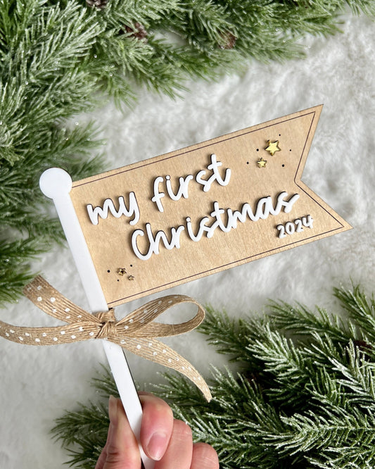 a flag made of birch wood and white acrylic that says my first christmas 2024 with gold stars around