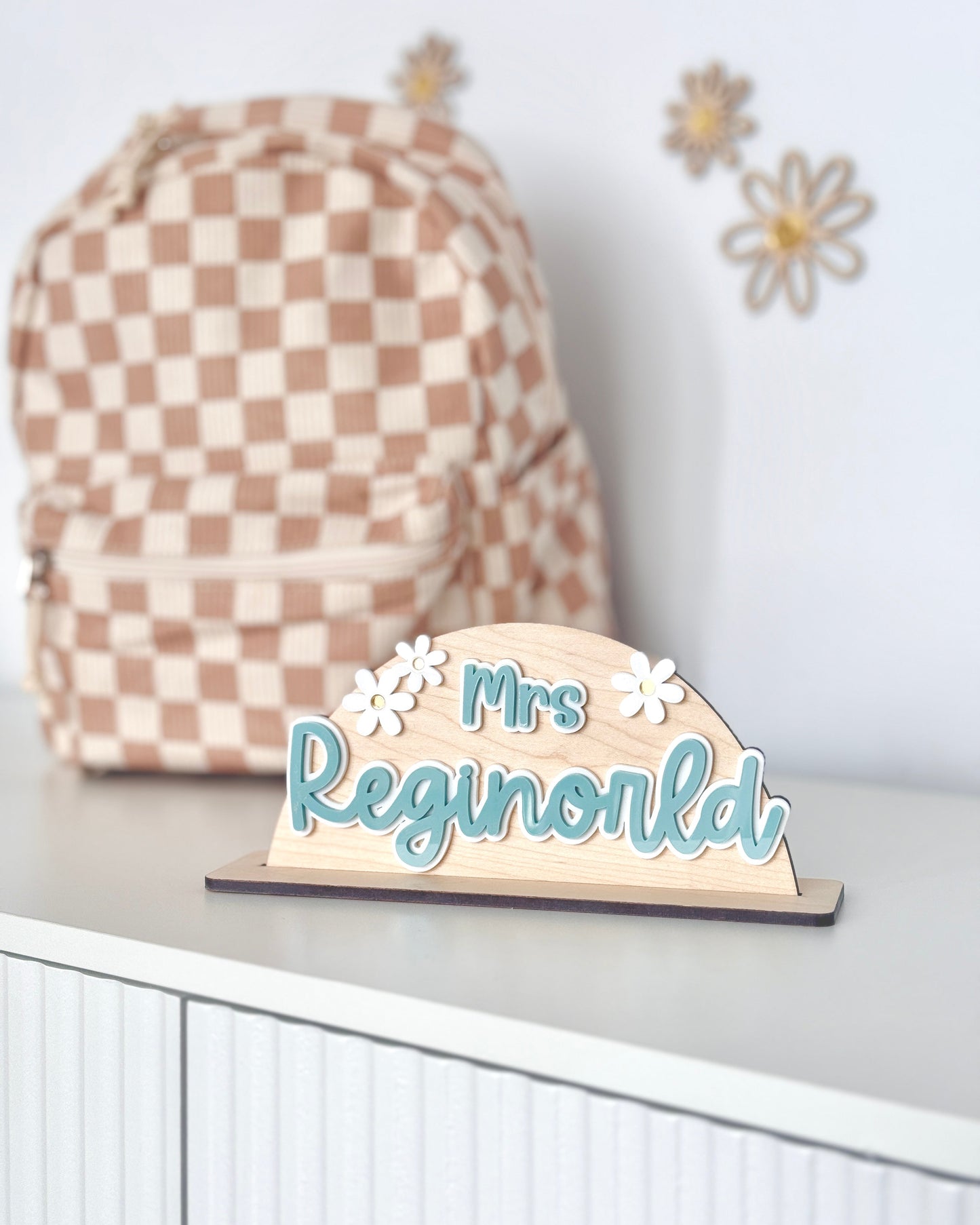Wooden nameplate with 'Mrs Reginworld' on a white surface with a checkered bag in the background.