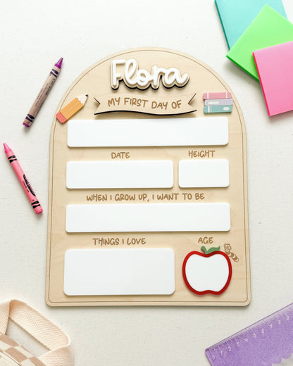 A wooden arch-shaped children's journal titled 'Flora My First Day of' laid flat on a white surface with colorful stationery props around it, including crayons, sticky notes, a ruler, and a small apple illustration.
