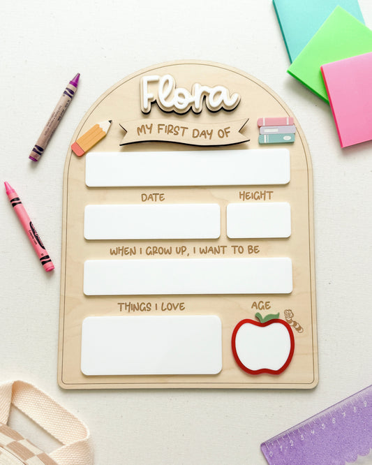 A wooden arch-shaped children's journal titled 'Flora My First Day of' laid flat on a white surface with colorful stationery props around it, including crayons, sticky notes, a ruler, and a small apple illustration.