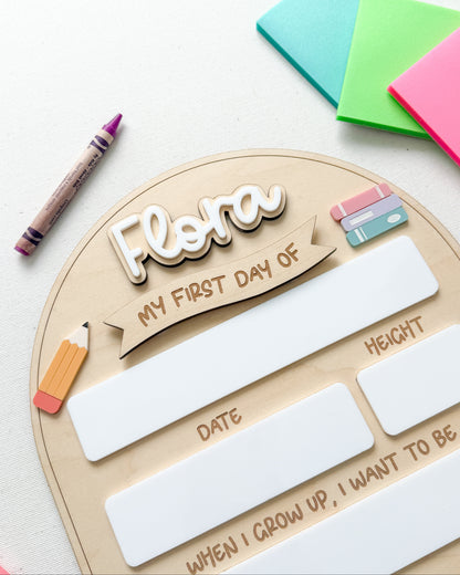 A wooden first-day-of-school milestone plaque with the name 'Flora' and writing spaces for date, height, and a future goal, styled on a white surface with colorful sticky notes, a crayon, and a pencil.