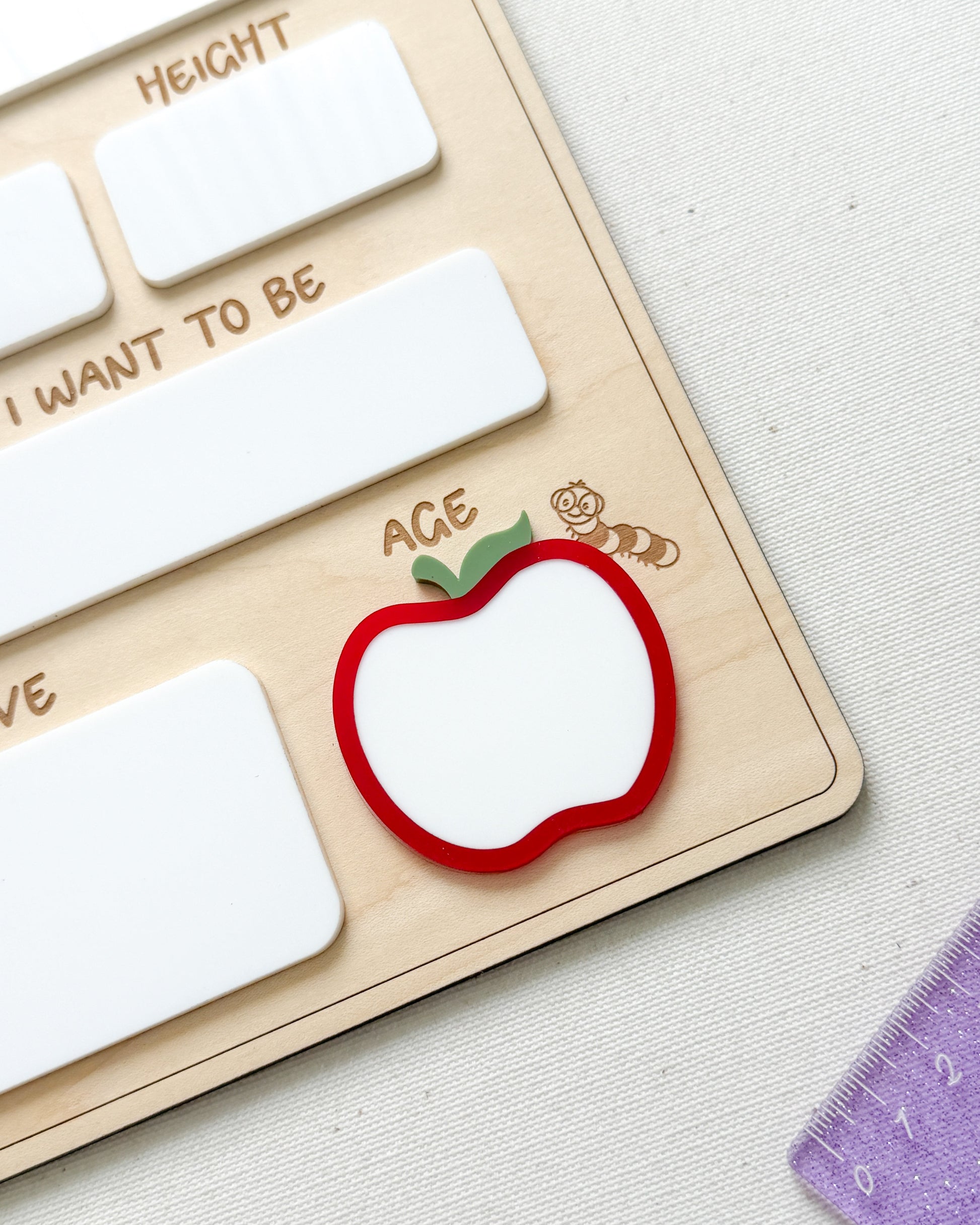 A close-up of a wooden Montessori-style educational board with white rectangular slots, engraved text, and a red apple-shaped piece with a green leaf and the word 'ACE' on a light fabric surface.
