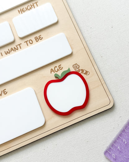 A close-up of a wooden Montessori-style educational board with white rectangular slots, engraved text, and a red apple-shaped piece with a green leaf and the word 'ACE' on a light fabric surface.