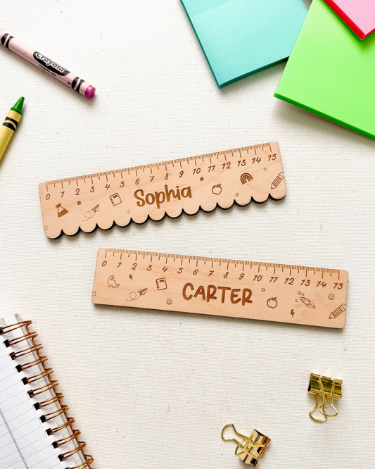Two personalized wooden school rulers with scalloped edges, engraved with names 'Sophia' and 'Carter' and ruler markings, arranged on a white desk with stationery props including sticky notes, crayons, binder clips, and a spiral notebook.