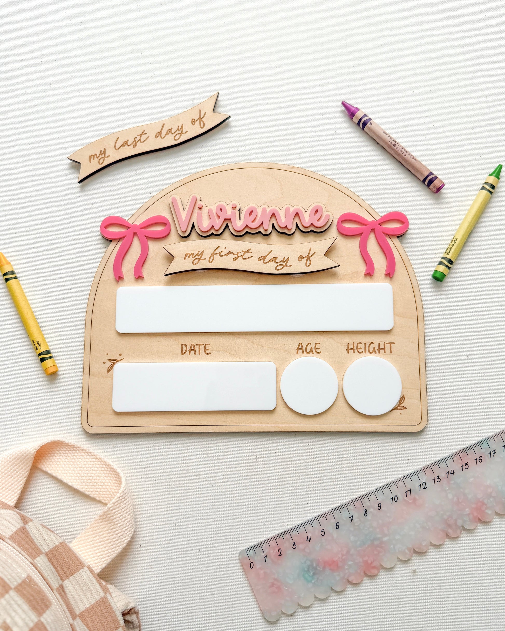 A personalized wooden first-day-of-school-supplies box with a pink bow and 'Vivienne my first day of' text, styled on a white background with crayons, a ruler, a checkered bag, and a paper banner.