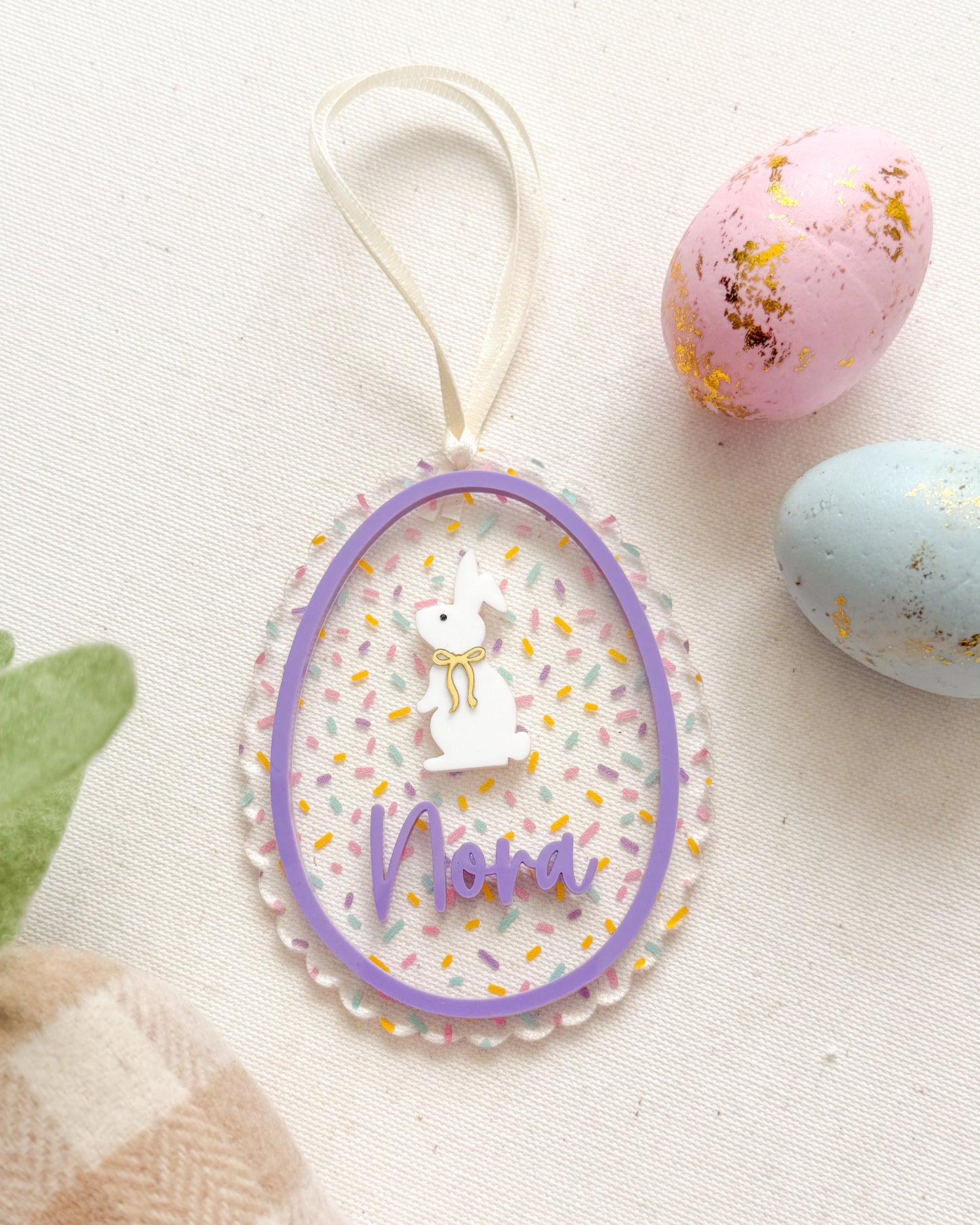 Decorative Easter egg with 'Nana' text, surrounded by pastel eggs and a checkered bag on a light background.