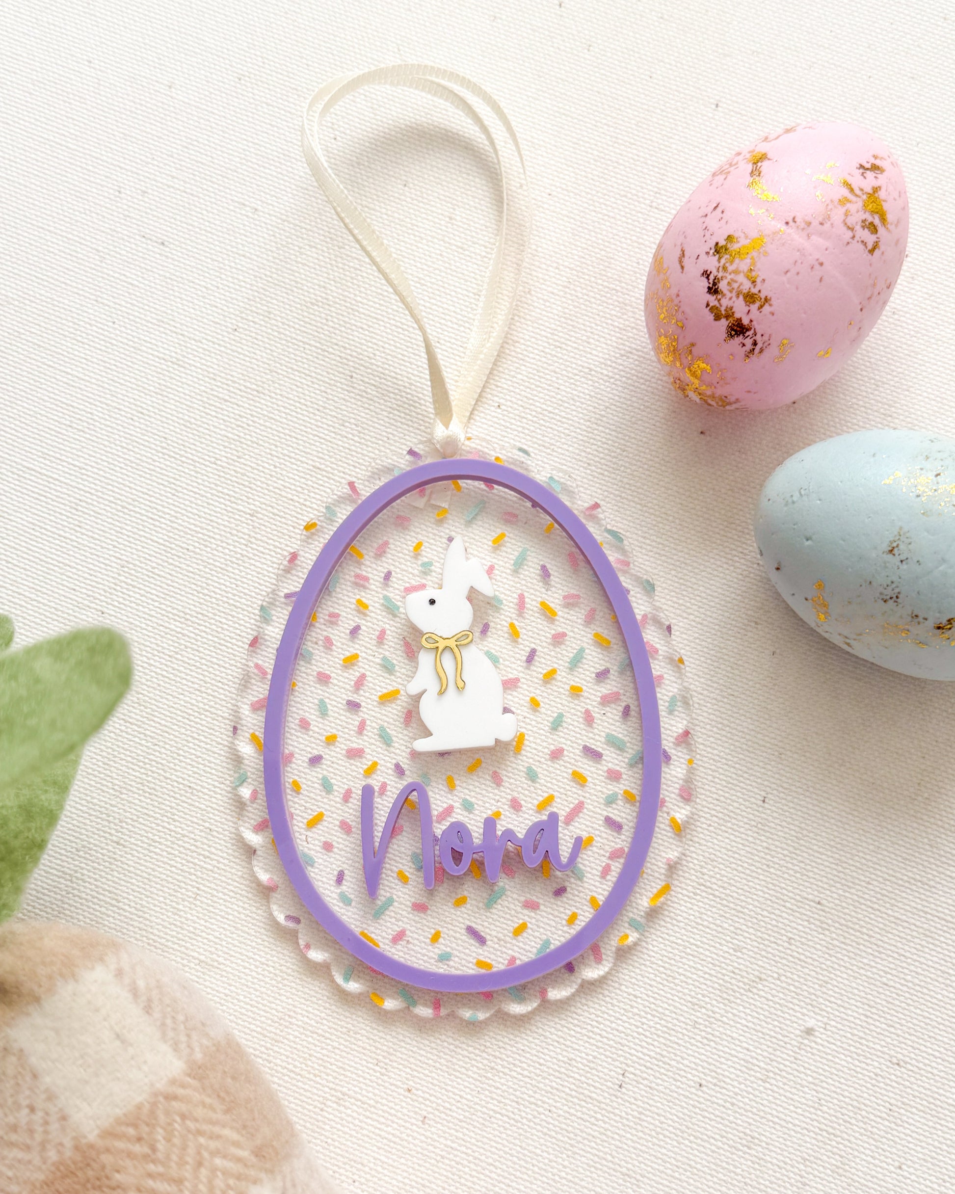 Decorative Easter egg with 'Nana' text, surrounded by pastel eggs and a checkered bag on a light background.