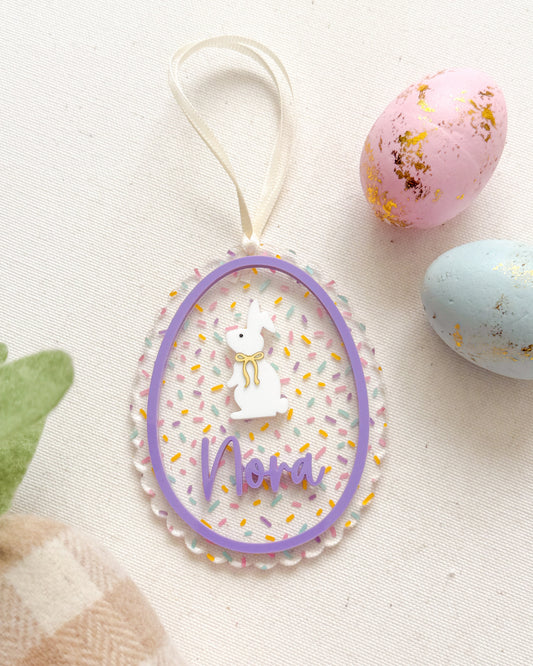 Decorative Easter egg with 'Nana' text, surrounded by pastel eggs and a checkered bag on a light background.