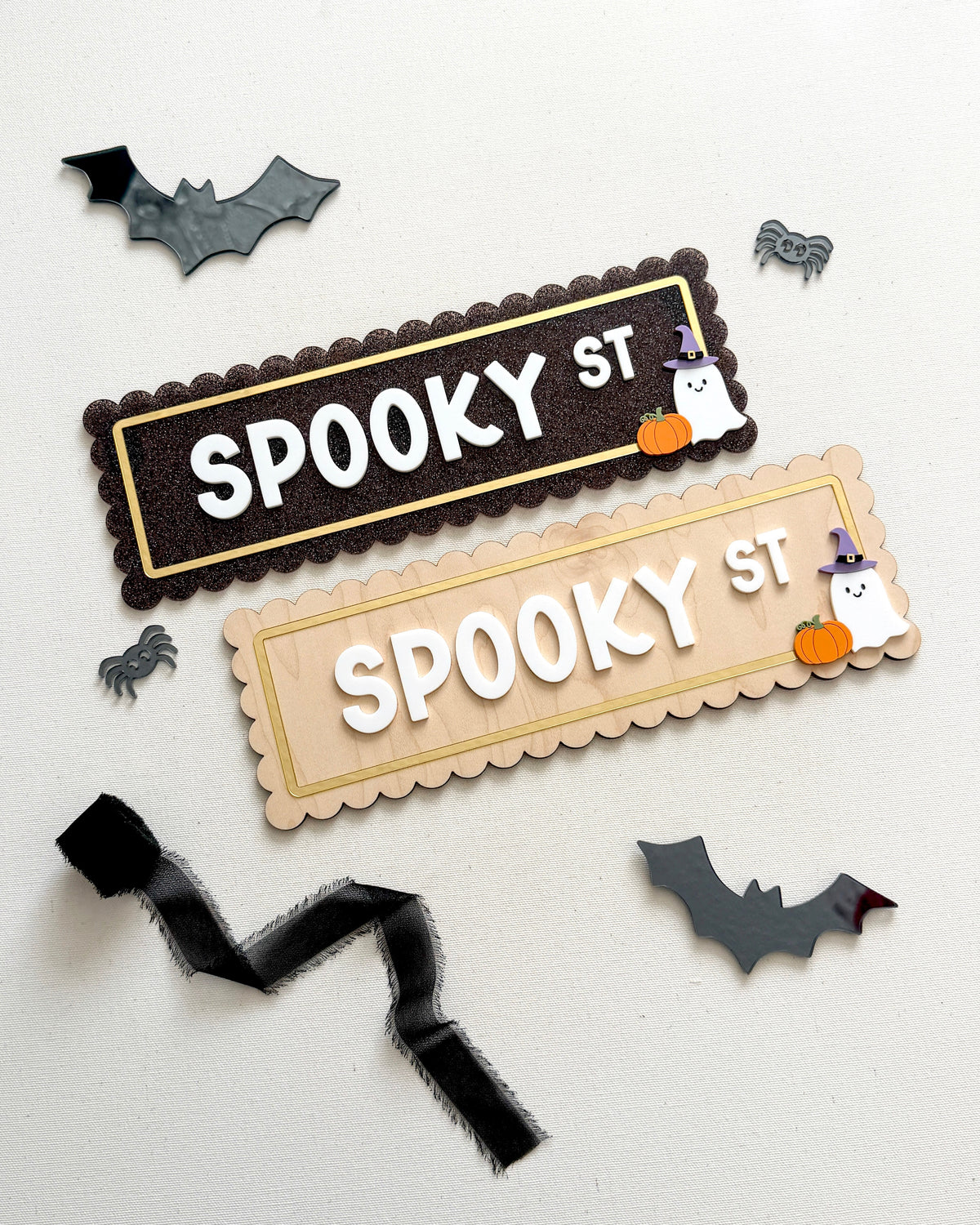 Two Halloween-themed decorative sign pieces reading “SPOOKY ST” on a white background, surrounded by small bat, spider, ghost, and pumpkin cutouts. One sign is dark brown with gold trim and white lettering, and the other is light beige with gold trim and white lettering.
