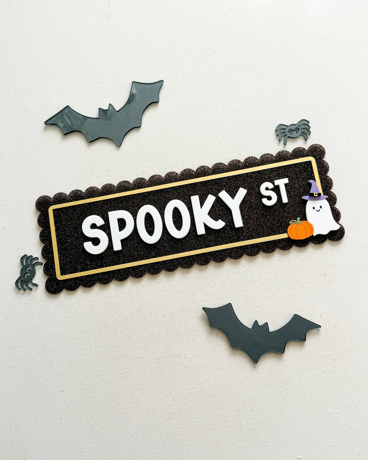A Halloween-themed decorative sign reading 'SPOOKY ST' in white letters on a black scalloped background with a gold inner border. The sign is styled with small bat, spider, ghost, and pumpkin cutouts on an off-white textured background.