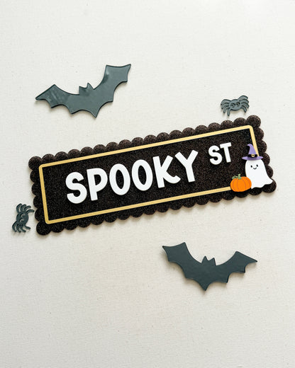A Halloween-themed decorative sign reading 'SPOOKY ST' in white letters on a black scalloped background with a gold inner border. The sign is styled with small bat, spider, ghost, and pumpkin cutouts on an off-white textured background.