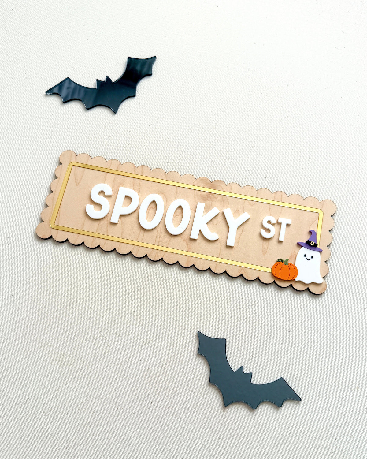 A scalloped wooden sign reading 'SPOOKY ST' in white letters, decorated with a small orange pumpkin, white ghost, and purple witch hat, displayed on a light surface with two dark bat cutouts nearby.
