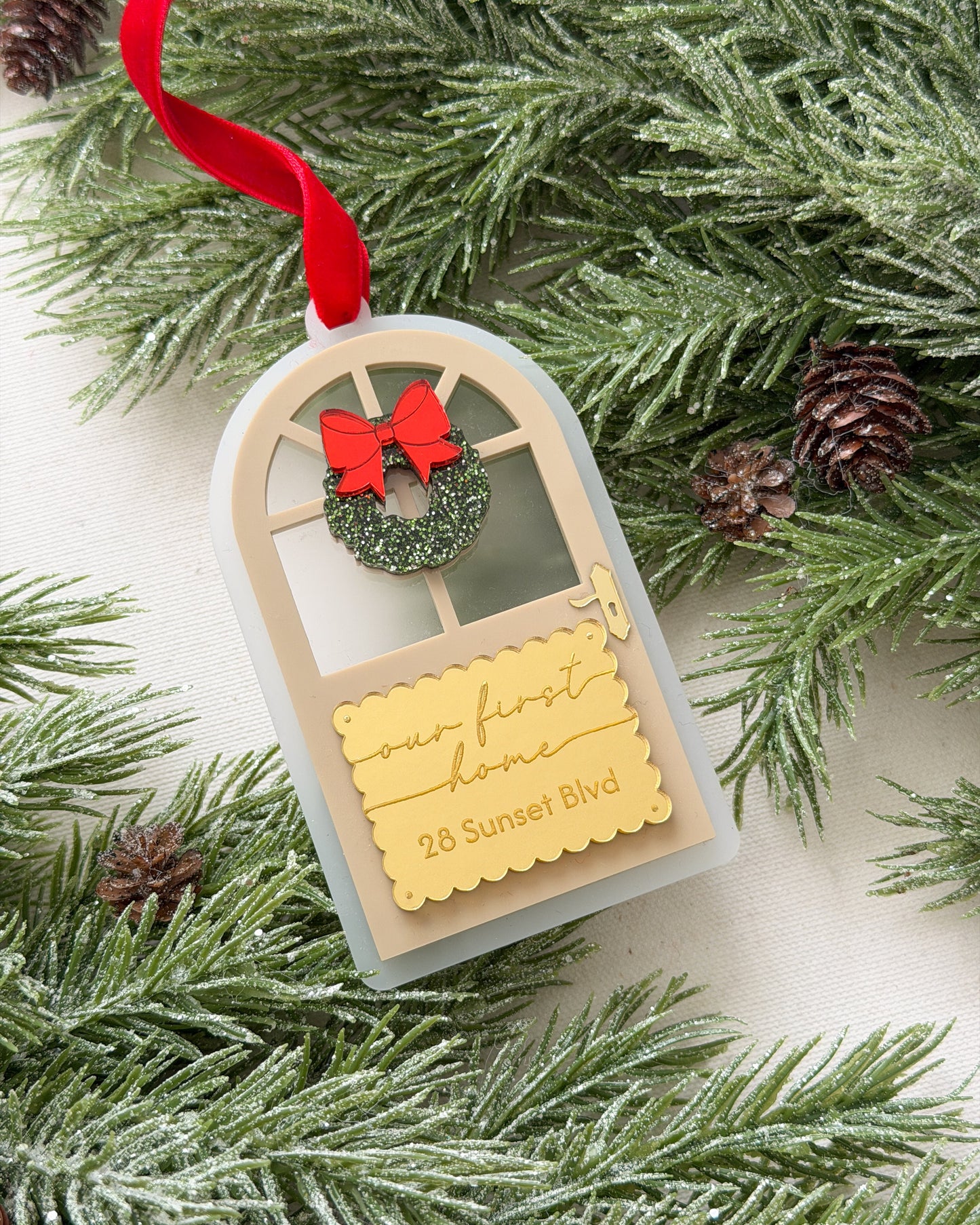 Decorative ornament with a red bow and wreath on a tag, surrounded by pine branches and cones.