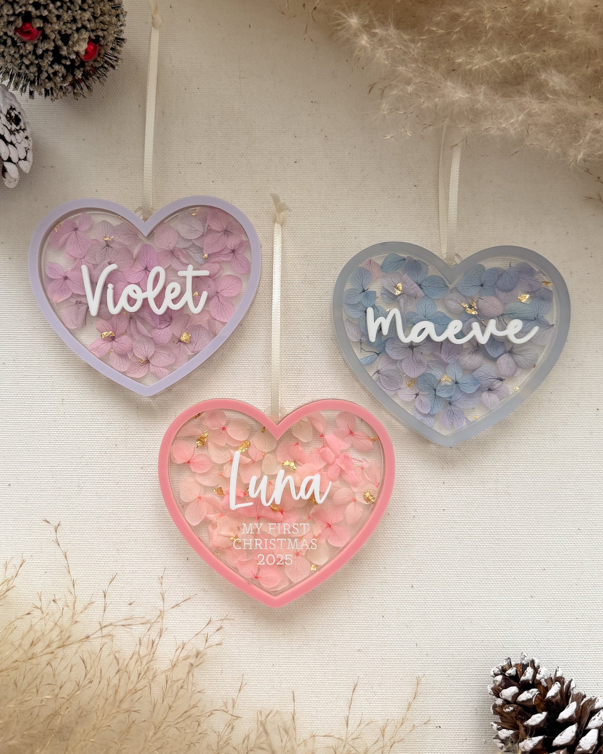 Heart-shaped name plaques with 'Violet', 'Maeve', and 'Luna' on a decorative background with pinecones and branches.