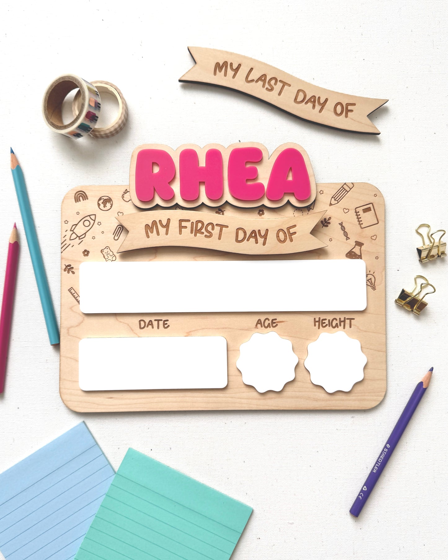 A wooden first-day-of-school milestone plaque with pink 'RHEA' lettering and blank white boxes for date, age, and height, styled on a white surface with colored pencils, sticky notes, washi tape, and paper clips.