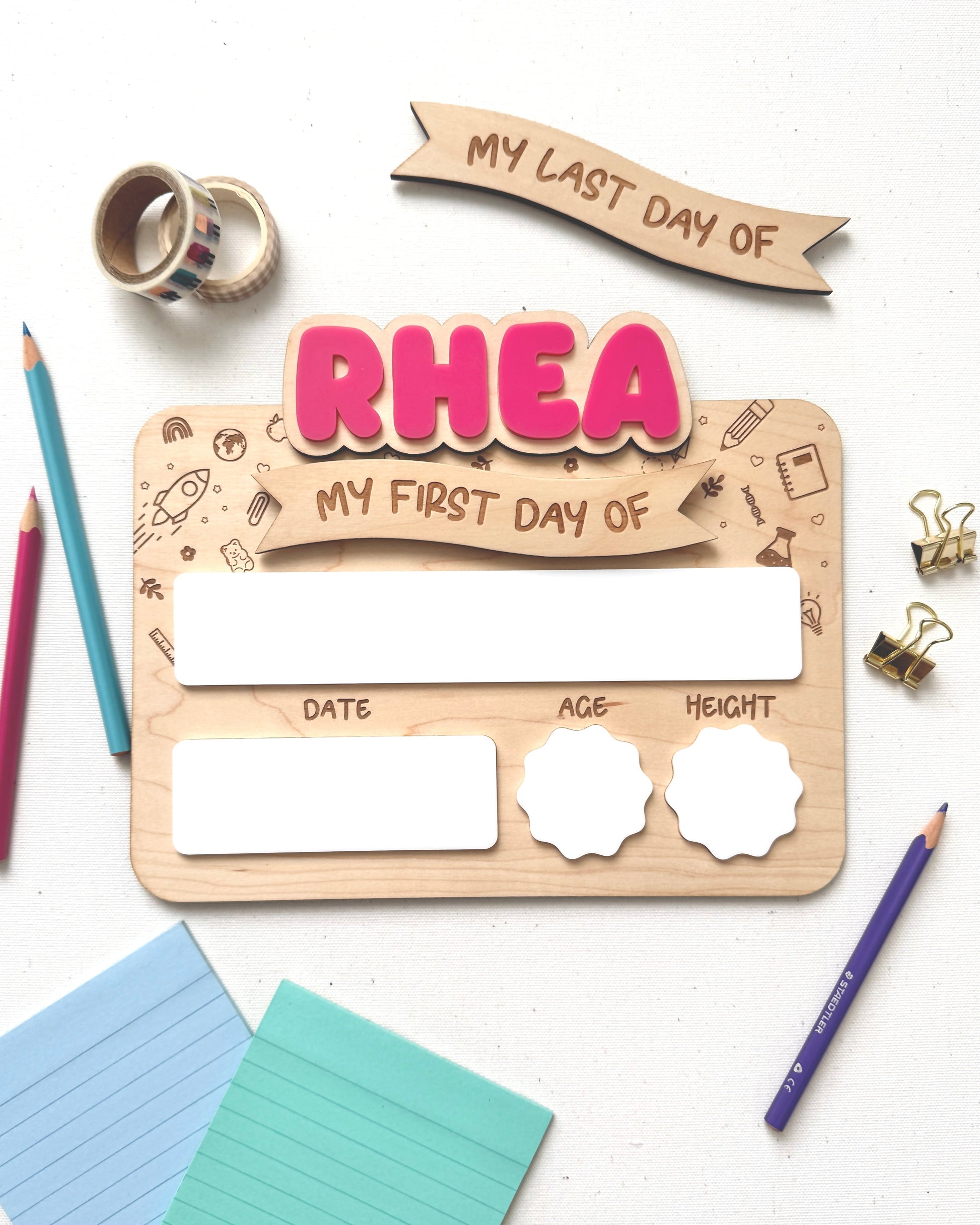 A wooden first-day-of-school milestone plaque with pink 'RHEA' lettering and blank white boxes for date, age, and height, styled on a white surface with colored pencils, sticky notes, washi tape, and paper clips.