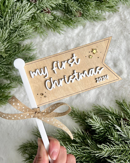 a flag made of birch wood and white acrylic that says my first christmas 2024 with gold stars around